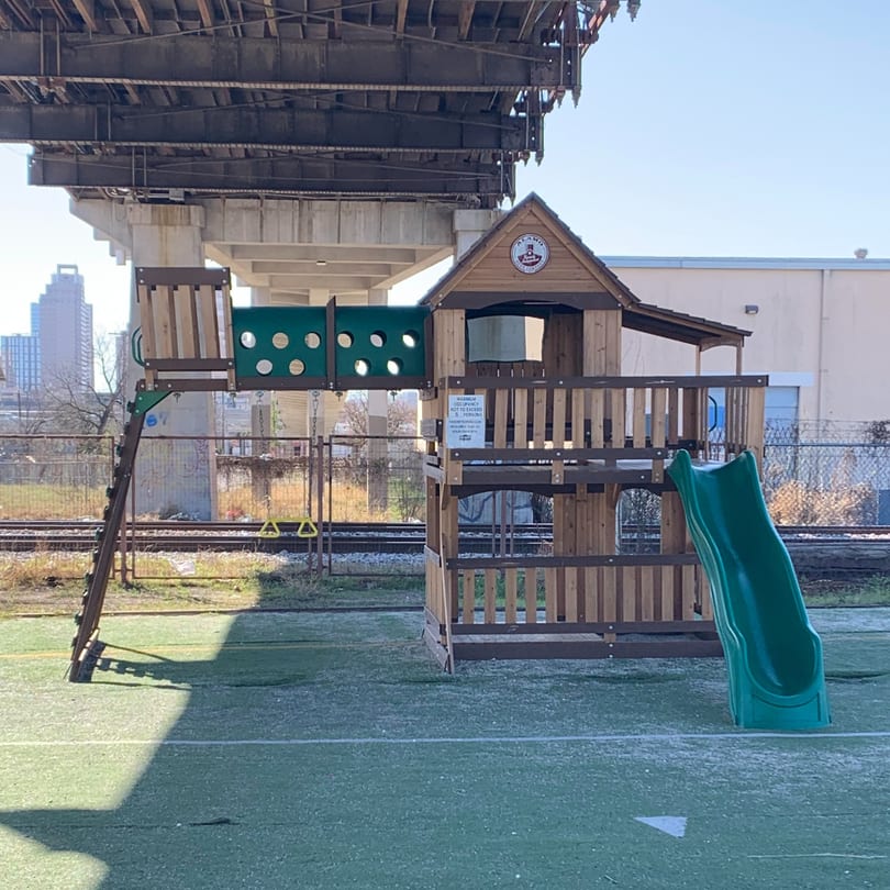 A wooden playground set with a green slide and climbing wall, under a large concrete overpass.