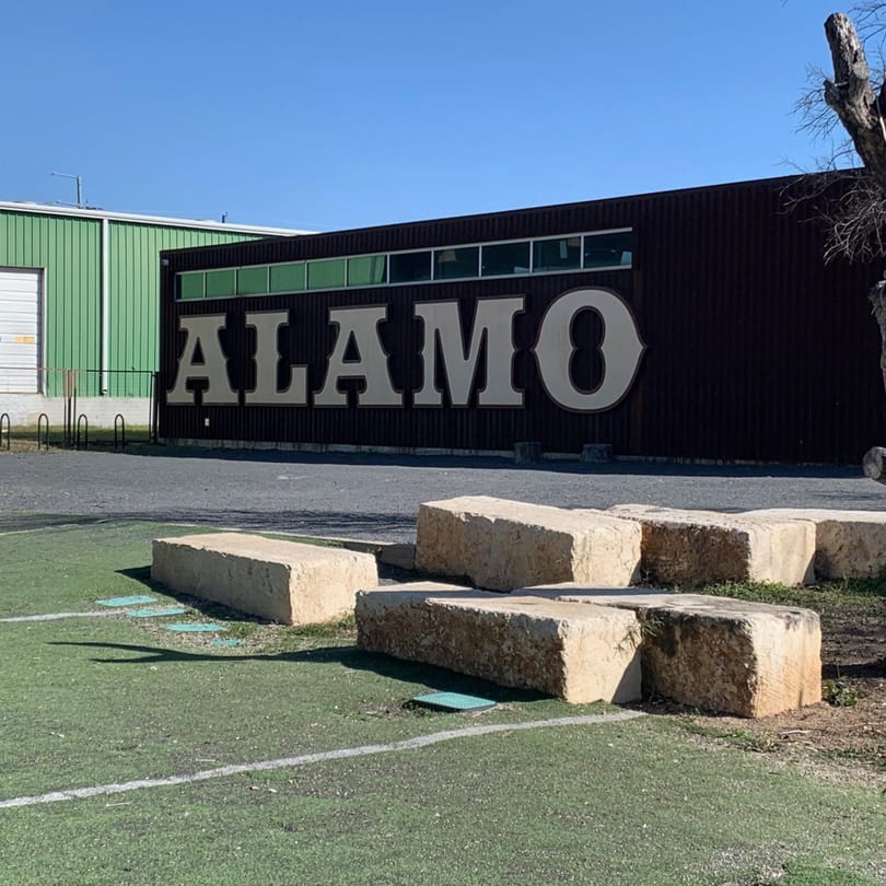 The word "ALAMO" in large white letters on a dark building, with green turf and stone blocks in the foreground.
