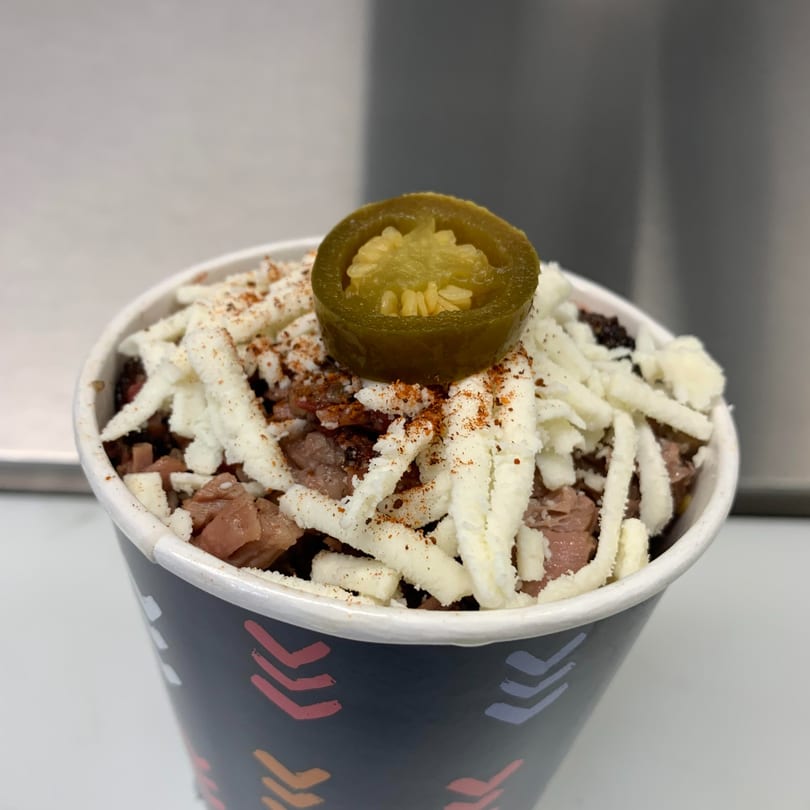 A Street Food Kings Mexican dish in a cup, topped with shredded cheese, spices, and a jalapeño slice.